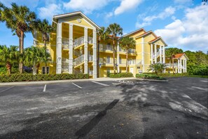 A yellow multi-story hotel with balconies, a parking lot, and palm trees.