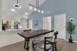 A spacious dining area with a long wooden table and chairs, a kitchen in the background, and a ceiling fan.