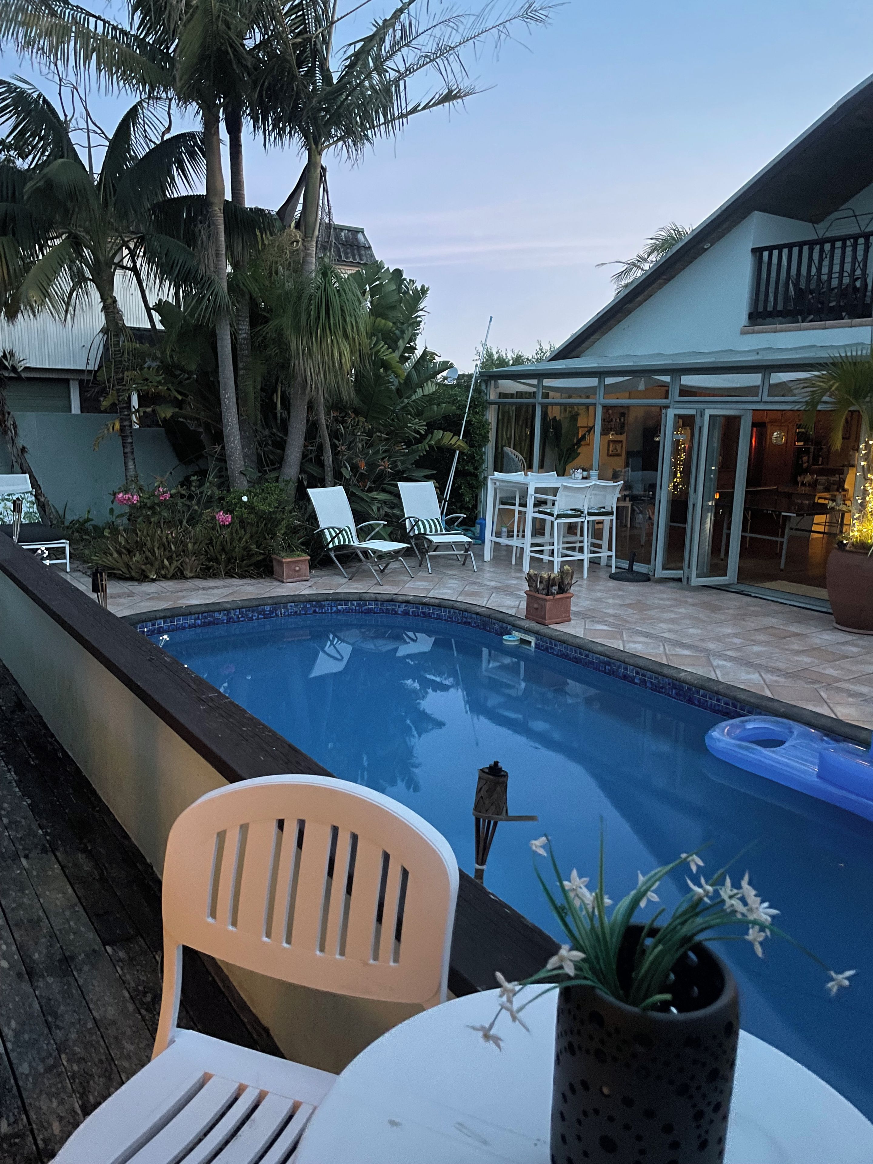 A swimming pool with lounge chairs and a table, surrounded by tropical plants and a house.