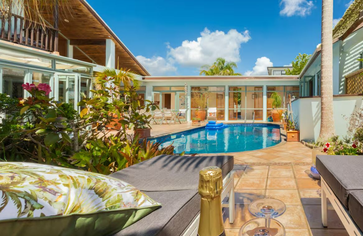 A pool area with lounge chairs and a hot tub, surrounded by tropical plants and a clear blue sky.