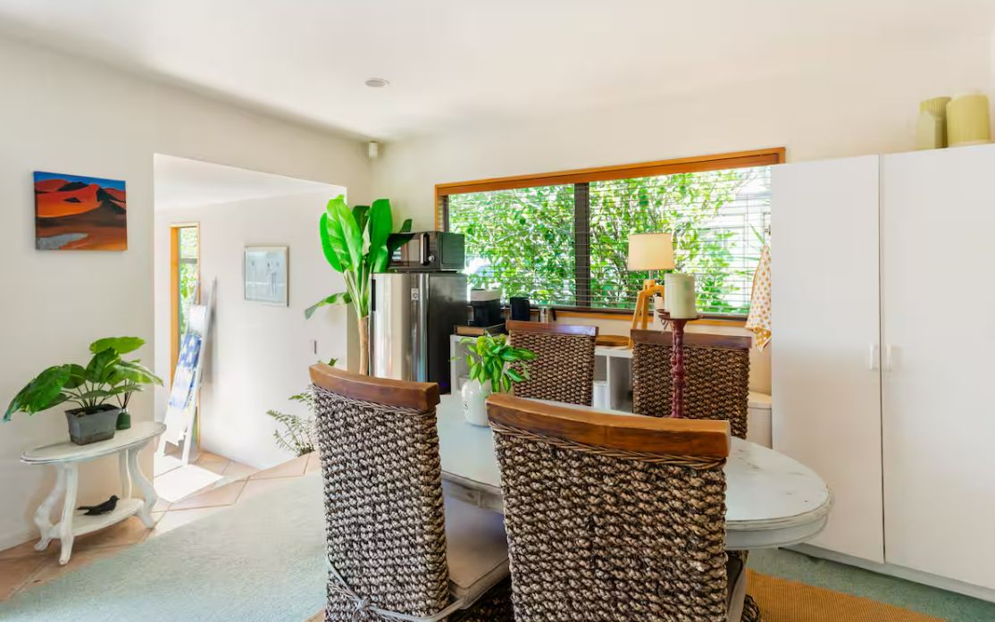 Un comedor con sillas de mimbre, una mesa redonda, un refrigerador y una ventana con vista a vegetación.