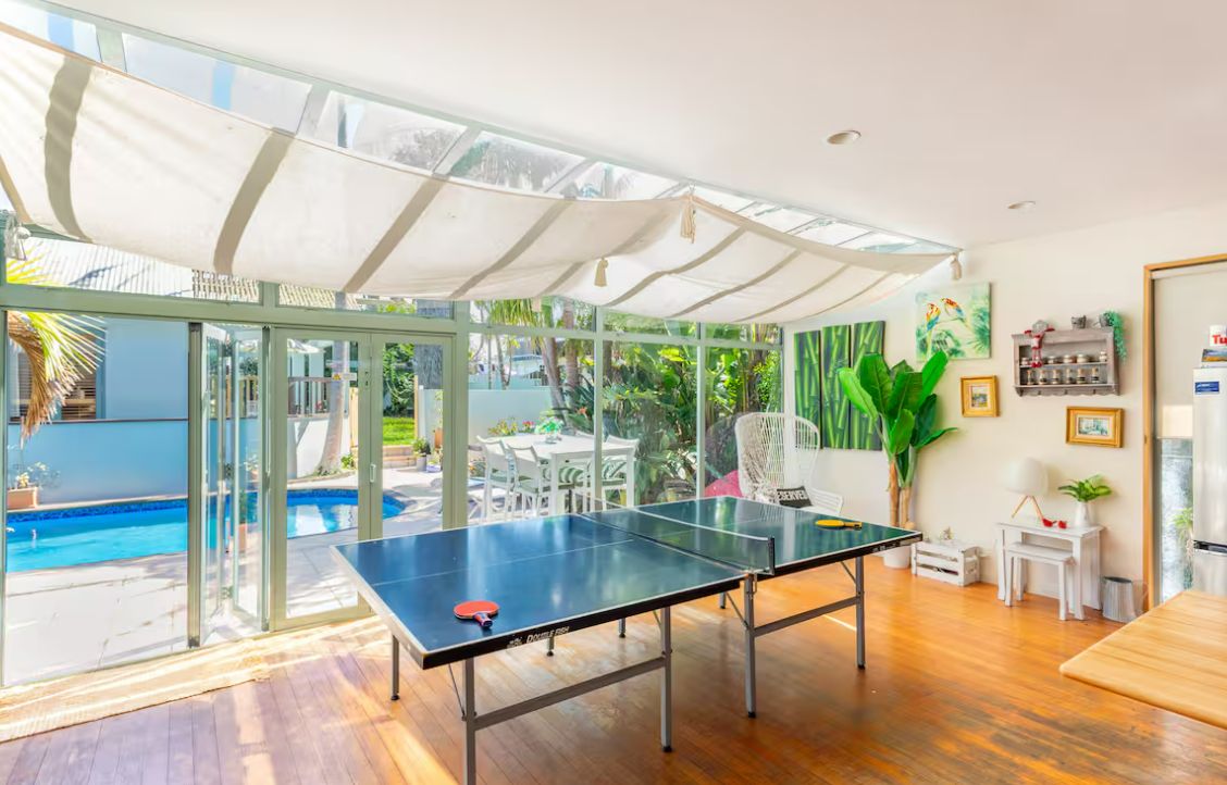 A ping pong table in a room with large windows, a pool, and a covered outdoor area.