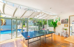 A ping pong table in a room with large windows, a pool, and a covered outdoor area.