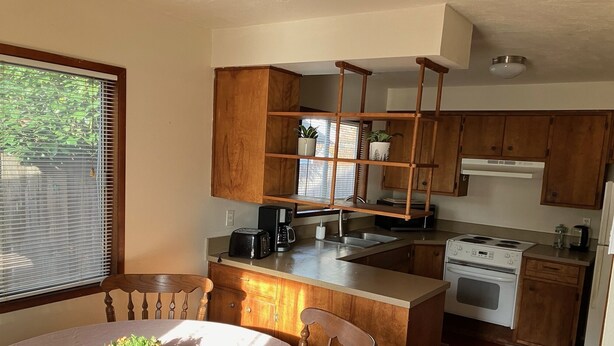 A compact kitchen with wooden cabinets, a built-in microwave, and a stove. There's a dining table with a plant centerpiece and a window with blinds.