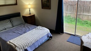 A bedroom with a bed, a nightstand with a lamp, a painting on the wall, and a view of the backyard through a sliding glass door.