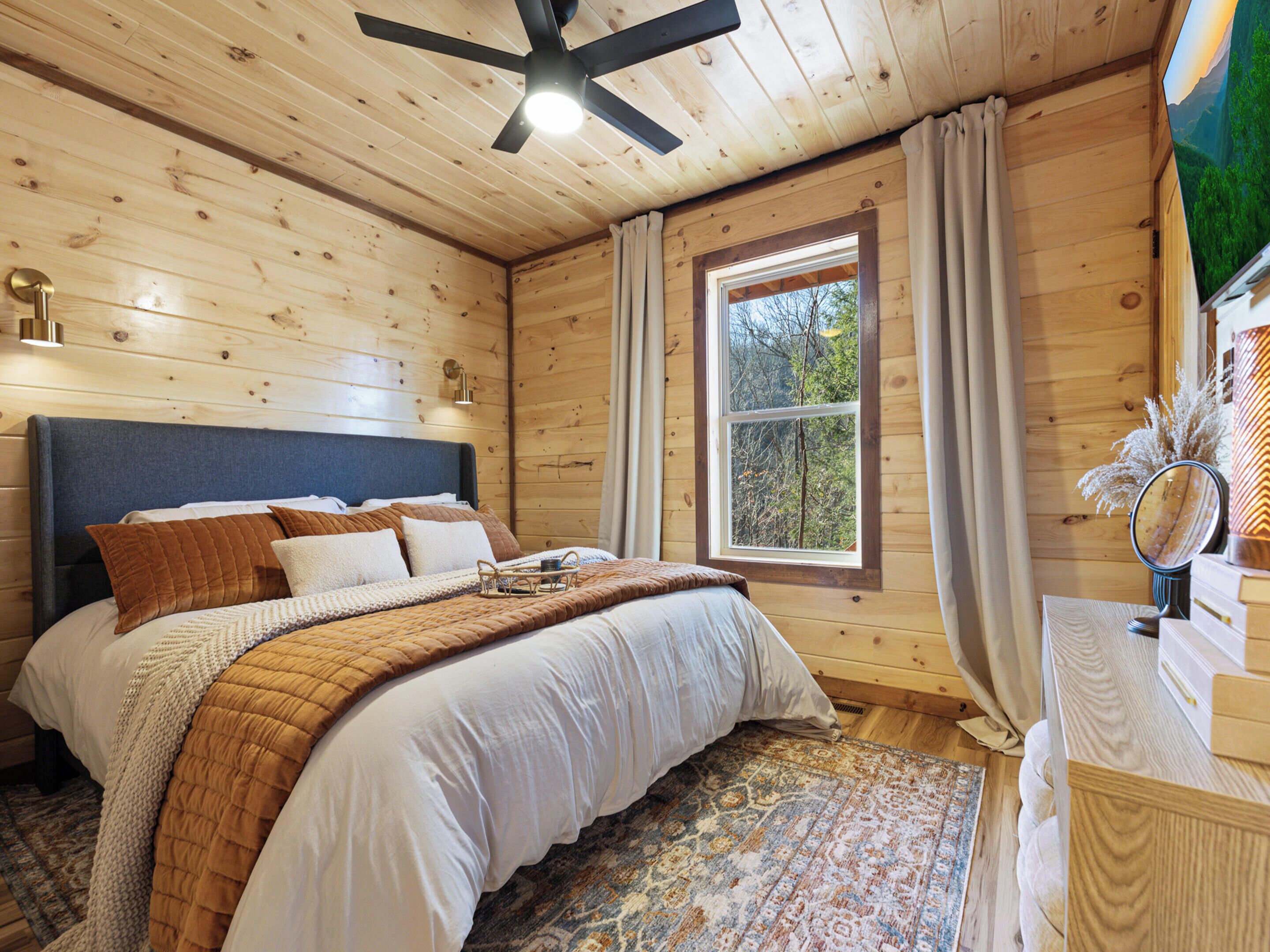 Un dormitorio de cabaña de madera con una cama grande, un ventilador de techo, una ventana con cortinas y un aparador de madera con espejo.