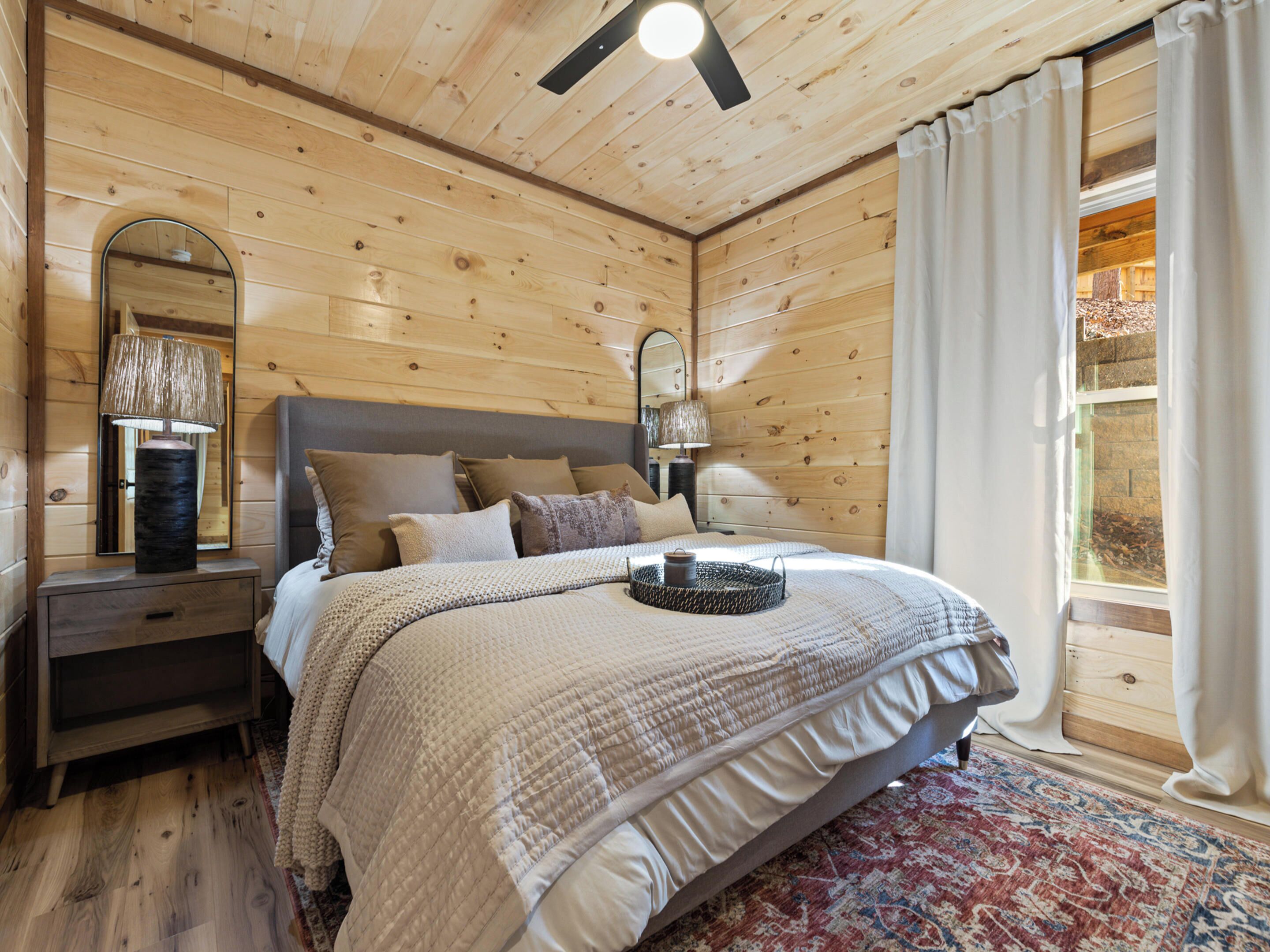 A wooden-paneled bedroom with a bed, nightstands, a mirror, and a window with curtains.