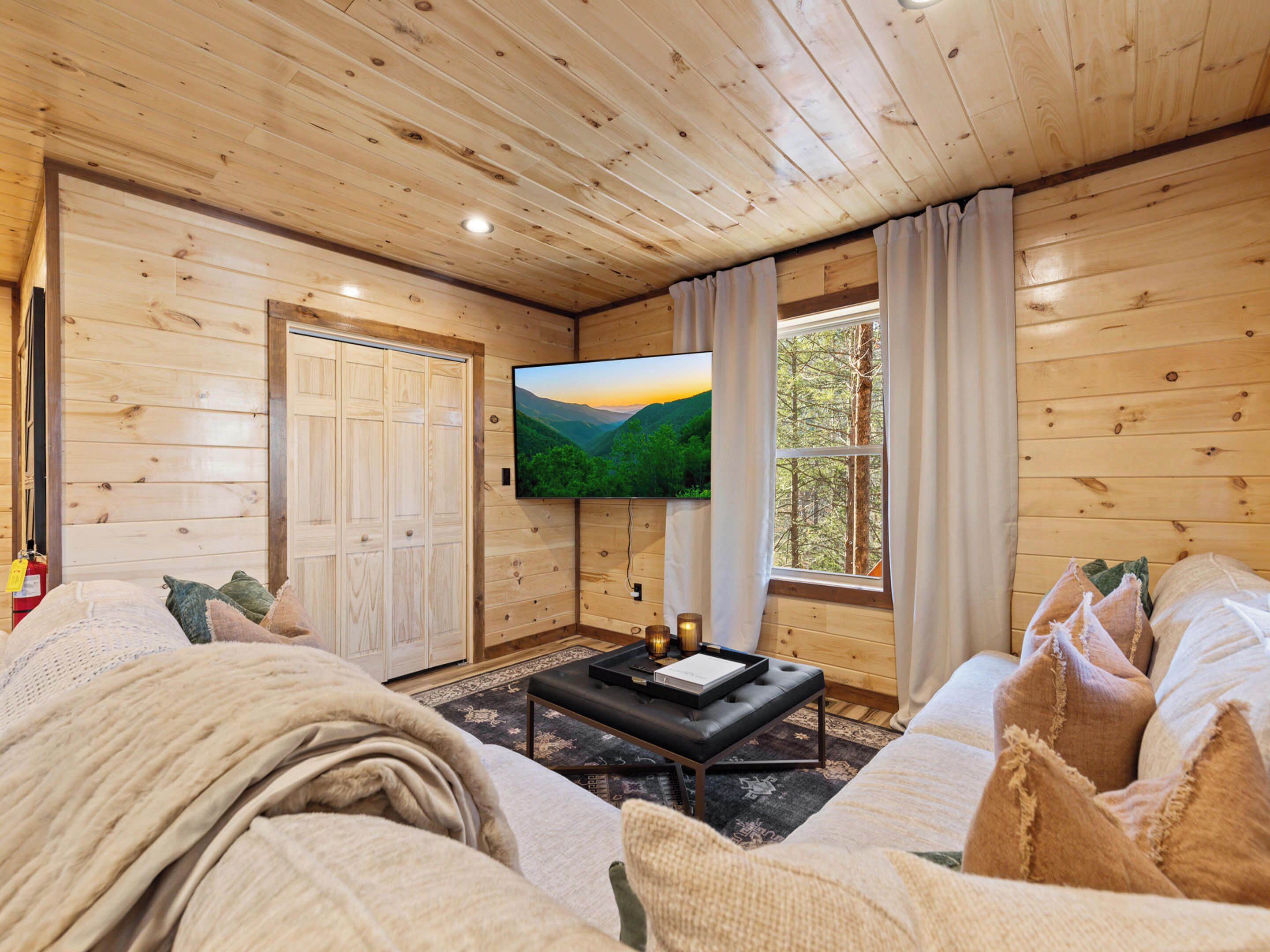 A cozy cabin interior with wooden walls, a large TV mounted on the wall, a comfortable sofa, and a window with curtains.