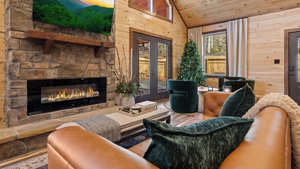 A cozy living room with a stone fireplace, a television mounted on the wall, a Christmas tree, leather sofas, and a wooden ceiling. - Mountain Ventures by Jackson Mountain Rentals (Pigeon Forge)