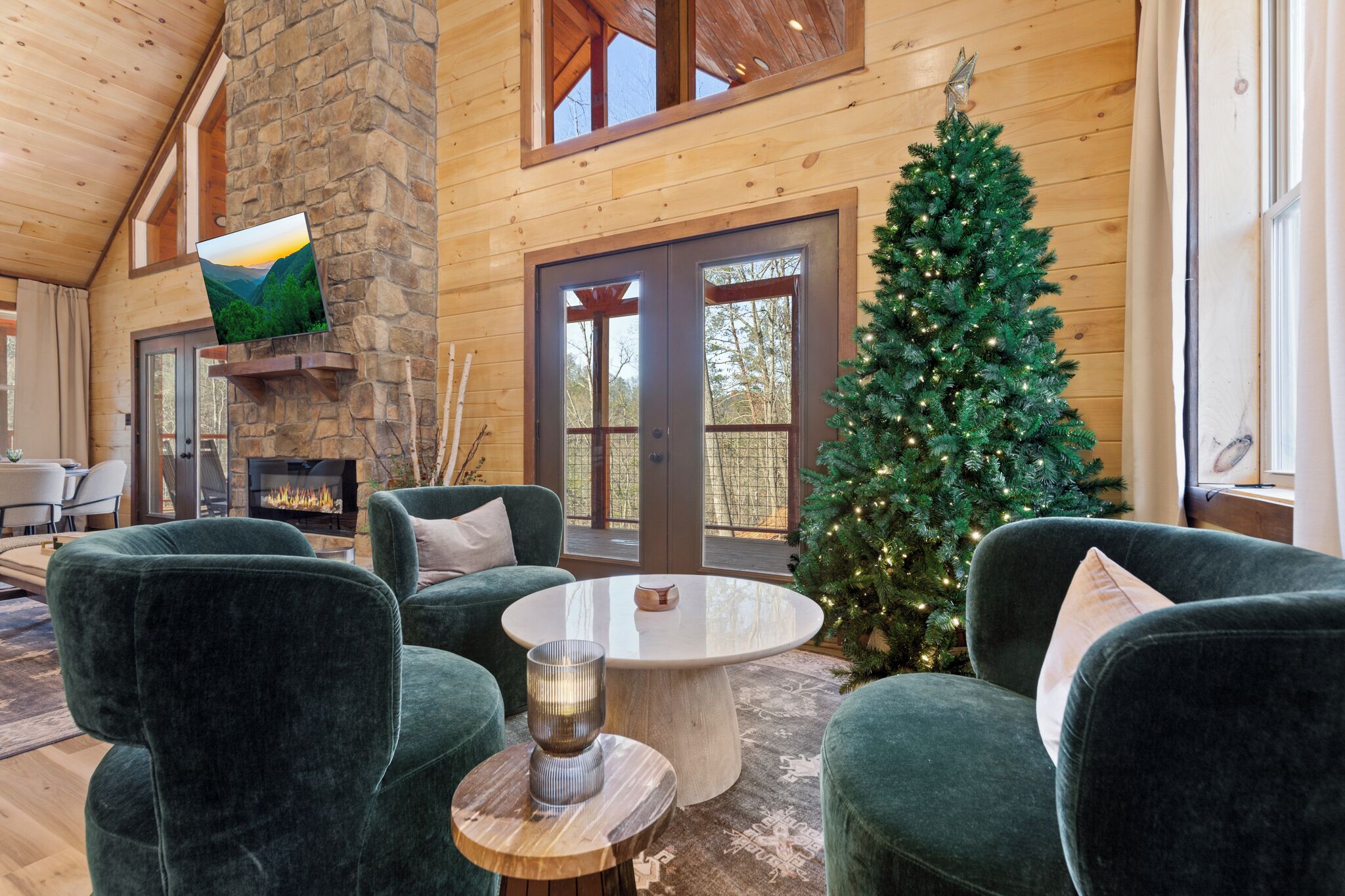 A cozy indoor setting with a stone fireplace, green velvet chairs, a Christmas tree, and a large window overlooking a forest.