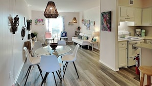 A modern living space with a dining area, a sofa, a kitchen, and a large window. - Let's Flamingle by Silver Sands Vacation Rentals (Port Aransas)