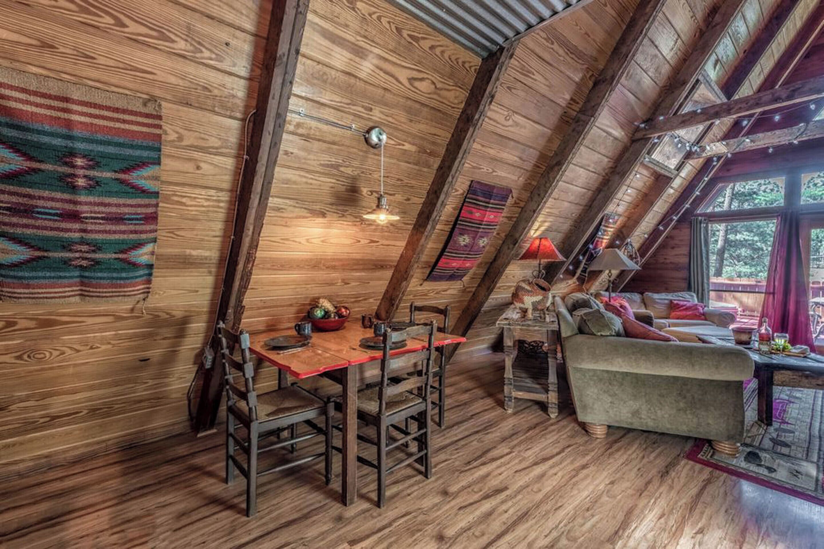 Um quarto de sótão acolhedor, com paredes de madeira, uma mesa de jantar com cadeiras, um sofá e uma janela com vista para árvores.