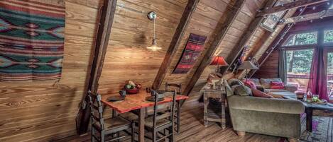 Um quarto de sótão acolhedor, com paredes de madeira, uma mesa de jantar com cadeiras, um sofá e uma janela com vista para árvores.