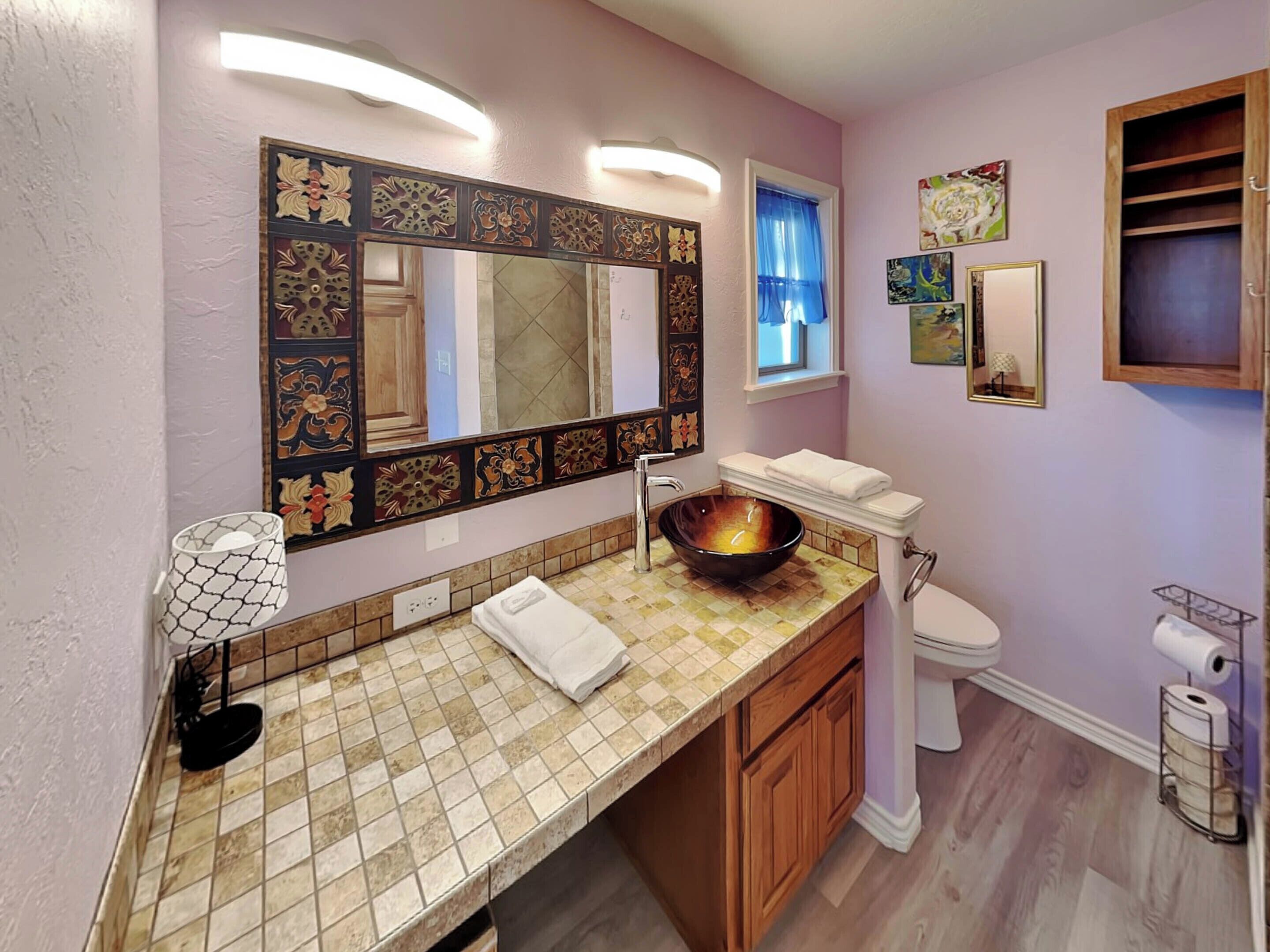 A bathroom with a tiled vanity, a large mirror, a vessel sink, a toilet, and a shelf with toiletries.