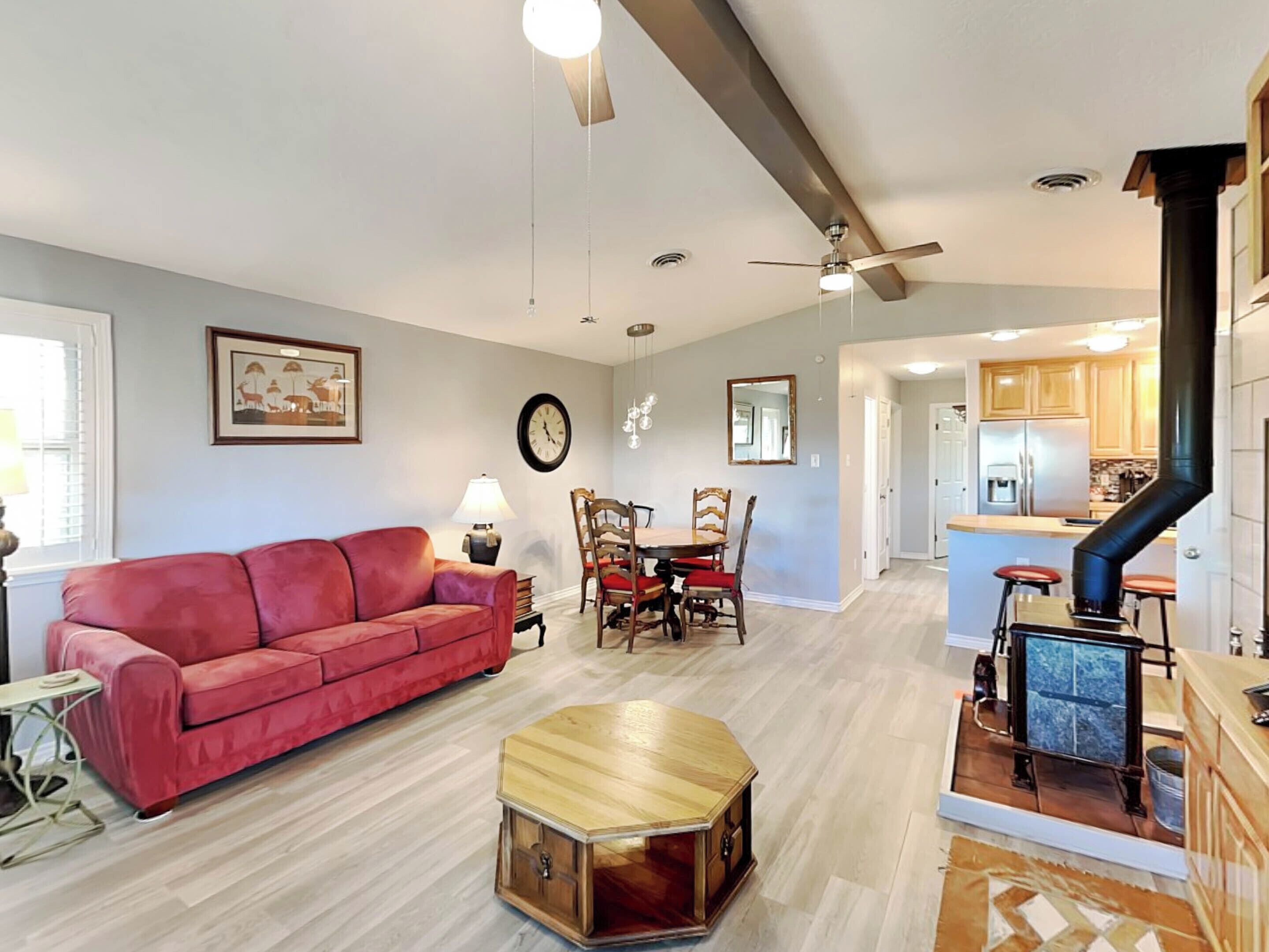 A living room with a red sofa, a wooden coffee table, a dining table with chairs, a clock, and a kitchen area with a stove and bar stools.