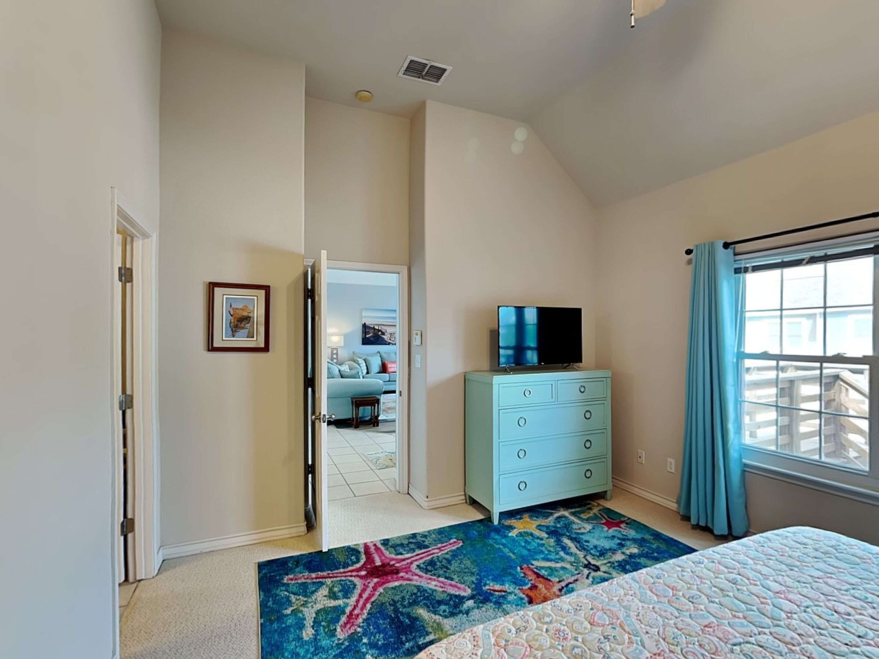 A bedroom with a bed, a dresser, a television on top, a starfish rug, and a view of a balcony through a window.