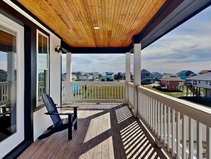 A covered porch with a wooden ceiling, a single black chair, and a view of a residential area with houses. - Pescador's Paradise by Ryson Vacation Rentals (Galveston)