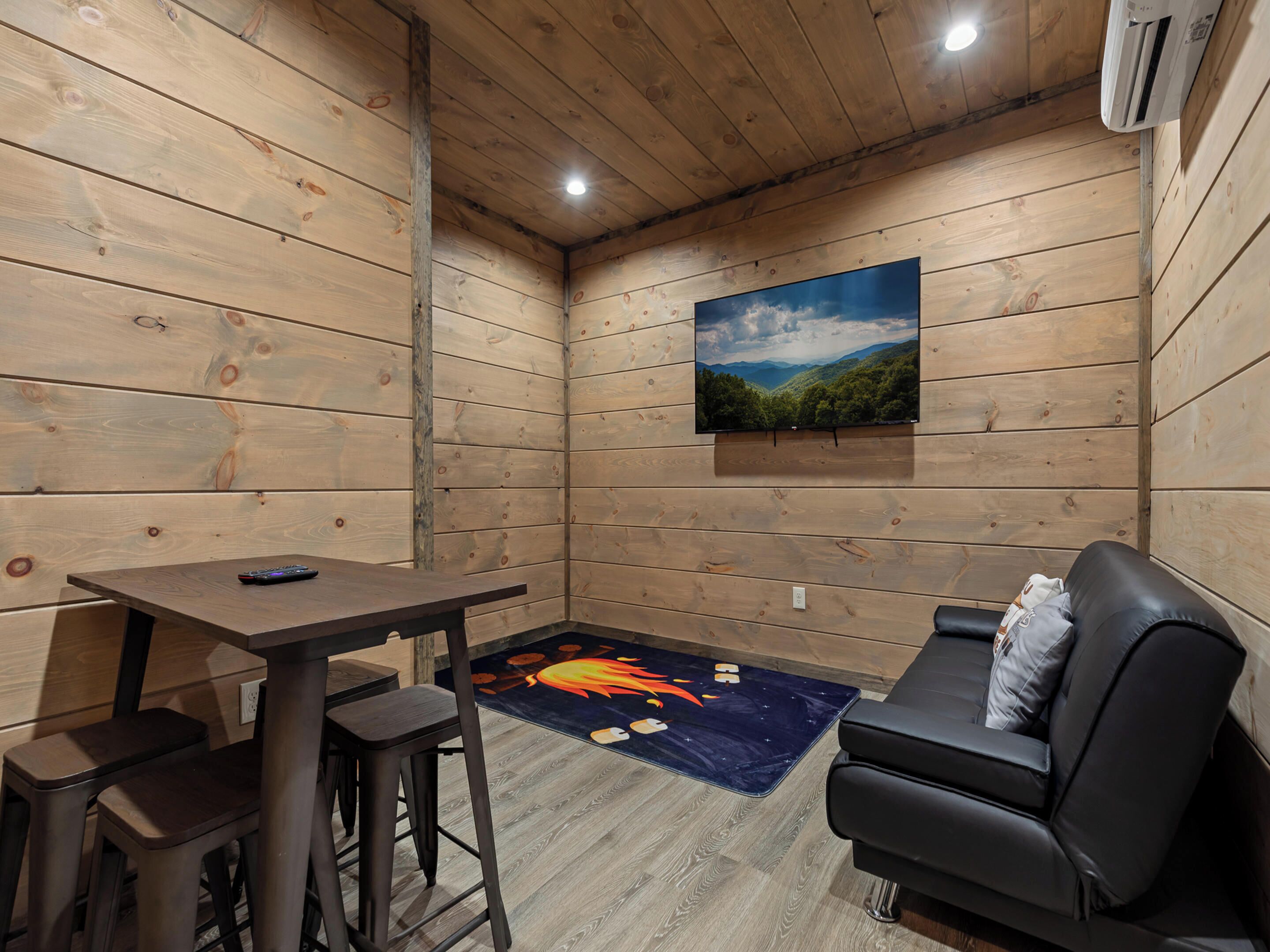 A cozy room with wooden walls, a mounted TV, a table with chairs, a sofa, and a rug with a mountain landscape.