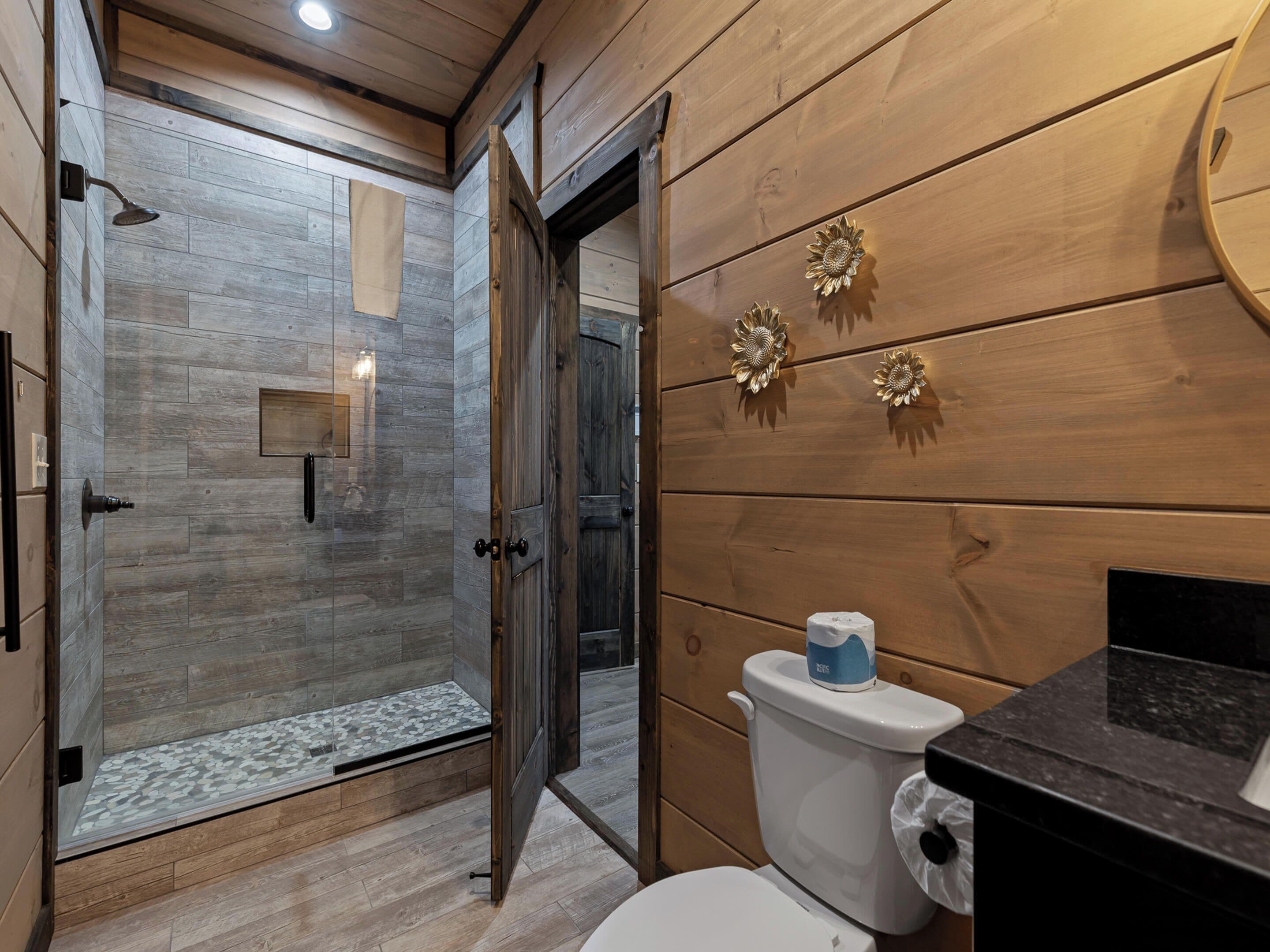 A modern bathroom with a glass shower, wooden walls, and a toilet with a paper towel holder.