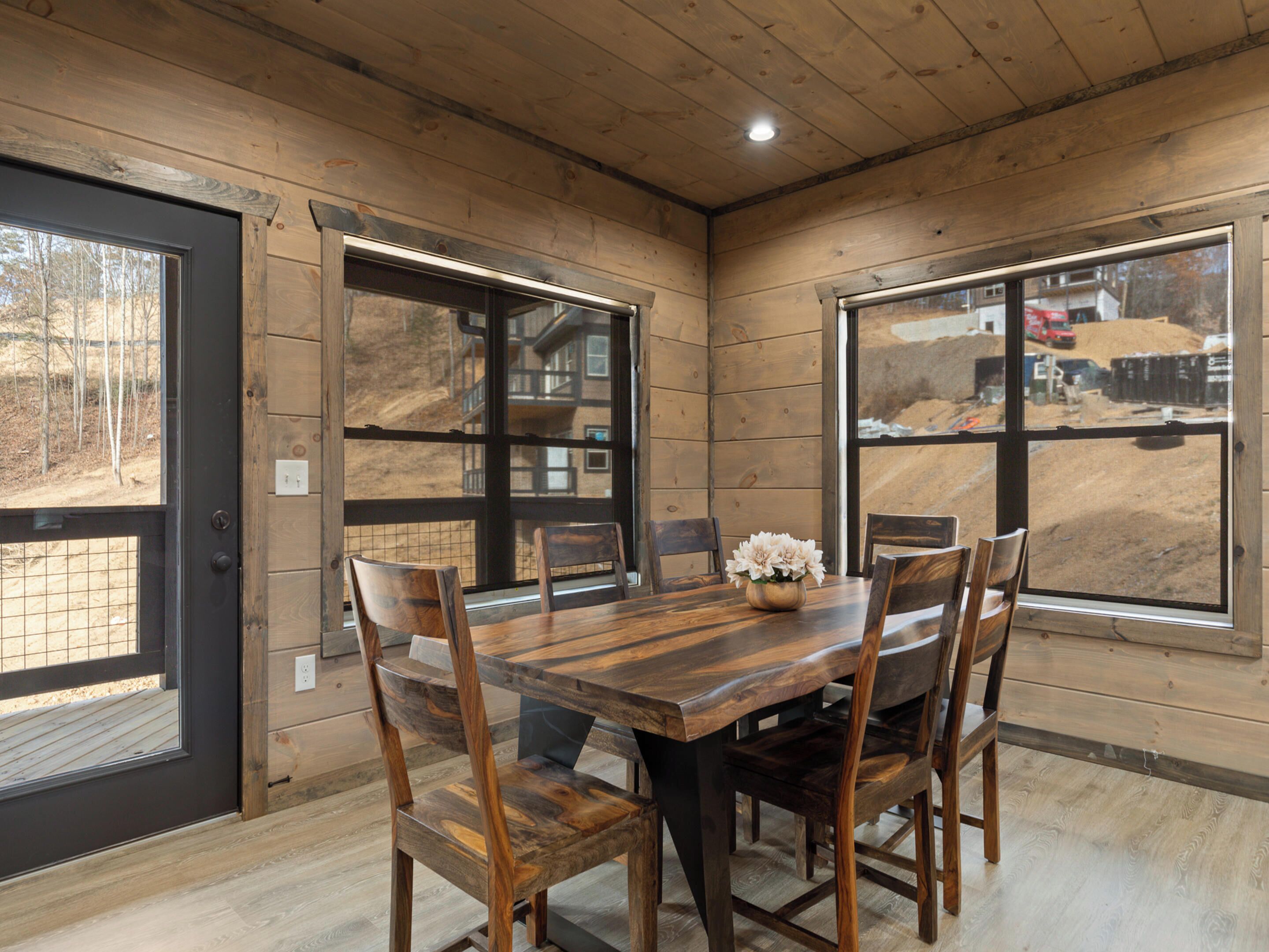 Un espace repas avec une table et des chaises en bois, de grandes fenêtres donnant sur un chantier de construction et un plafond en bois.