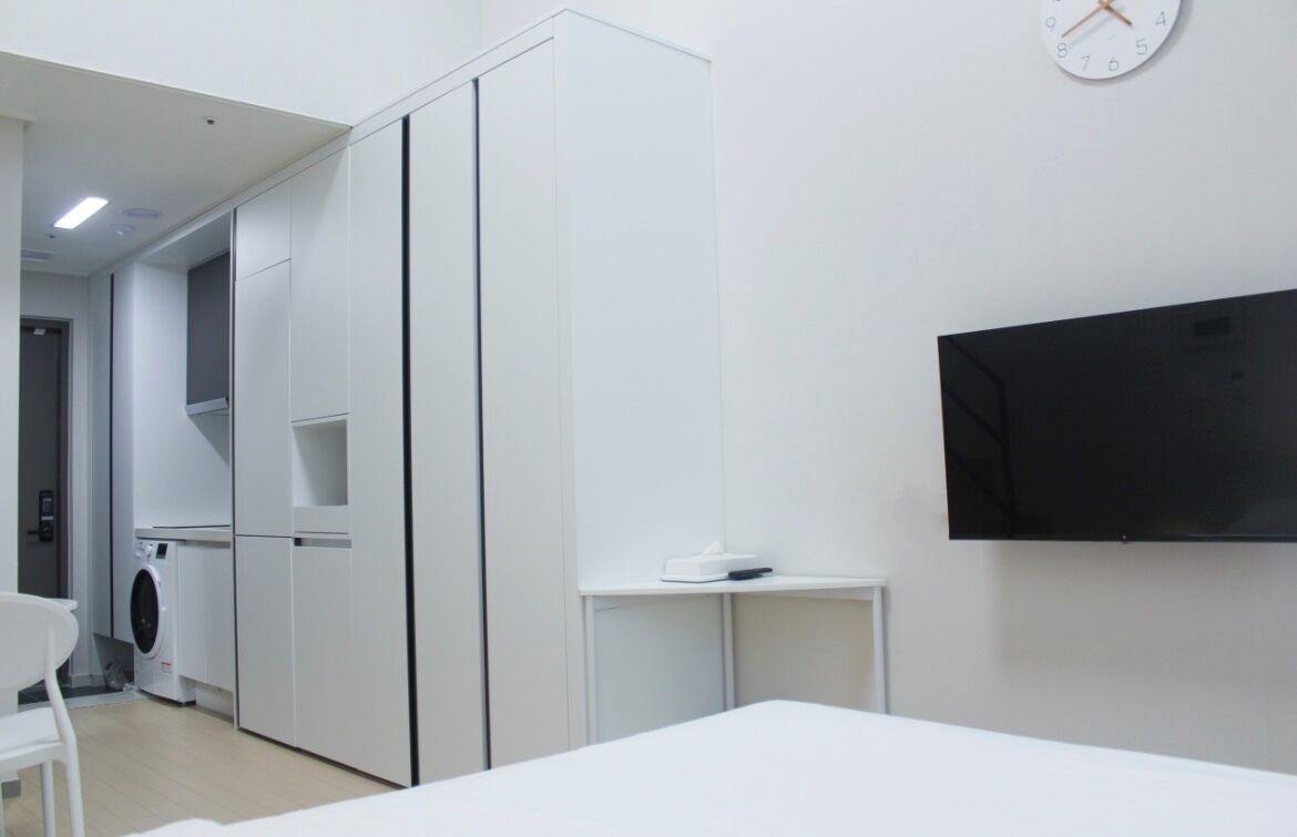 A modern hotel room with a bed, a TV mounted on the wall, a washing machine, and a small table with a scale.