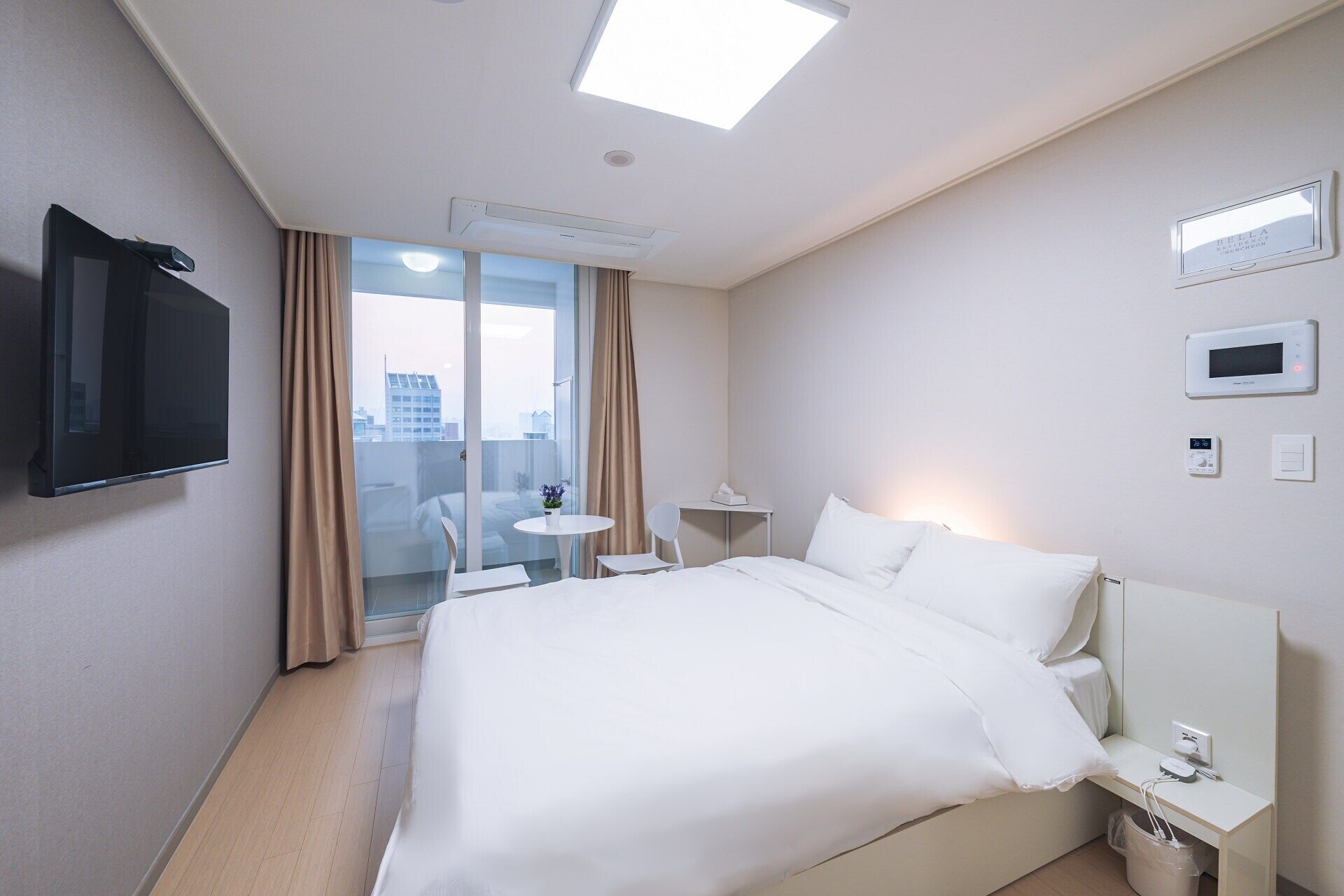Habitación de hotel con cama, televisión montada en la pared, una mesita con silla y ventana con cortinas.
