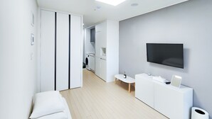 A modern, minimalist hotel room with a white wardrobe, a small seating area, a TV mounted on the wall, and a trash bin. - Chuncheon Bella Residence Hotel (Chuncheon)