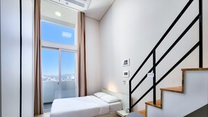 A modern bedroom with a bed, a chair, a small table, a staircase, and a city view through a large window. - Chuncheon Bella Residence Hotel (Chuncheon)