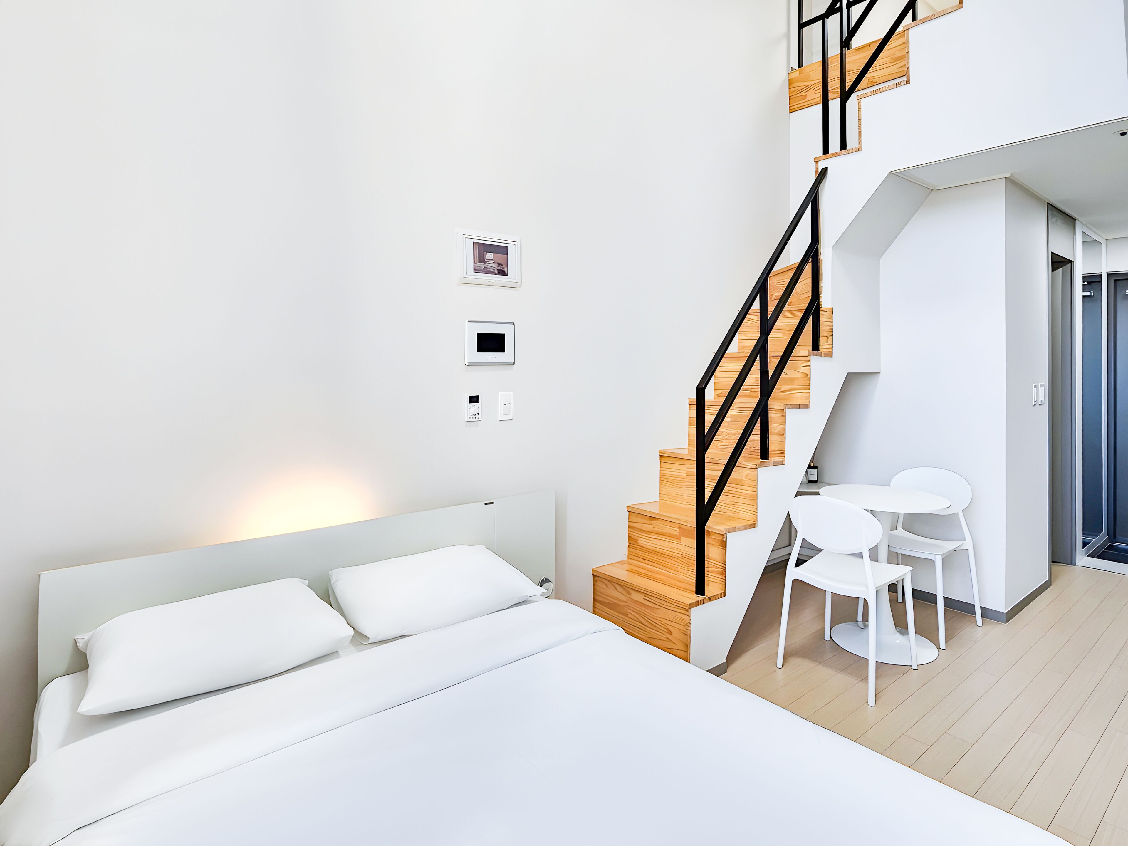 Un dormitorio moderno con una escalera de madera, una cama con ropa blanca, una pequeña mesa con sillas y un termostato fijado a la pared.