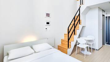 Un dormitorio moderno con una escalera de madera, una cama con ropa blanca, una pequeña mesa con sillas y un termostato fijado a la pared.