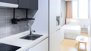 A modern kitchen with a built-in stove, a sink, and a black hanging rack. Adjacent to the kitchen is a bedroom with a bed, a bedside table, and a window with curtains.