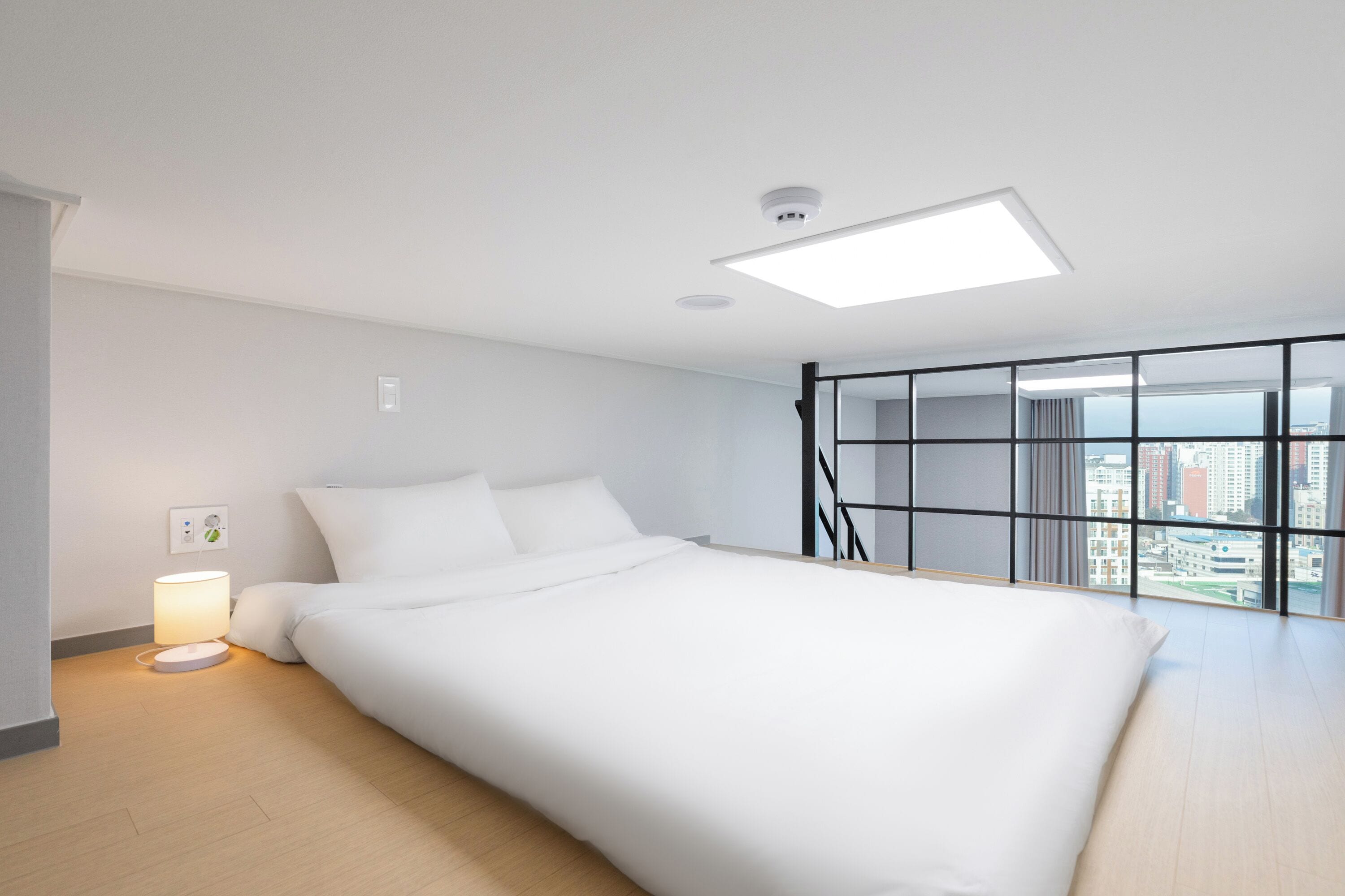 Een moderne hotelkamer met een groot bed, een nachtkastje met lamp, uitzicht op de stad door grote ramen en een plafondlamp.
