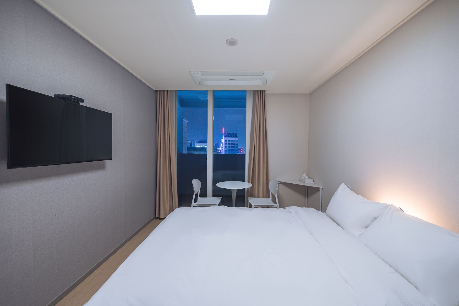 A hotel room with a bed, a TV mounted on the wall, a small table and chair, and a window with curtains.
