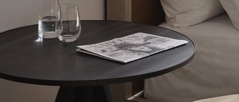 Une petite table ronde avec deux verres et une bouteille, un magazine et un canapé avec des coussins, le tout dans une chambre d’hôtel.