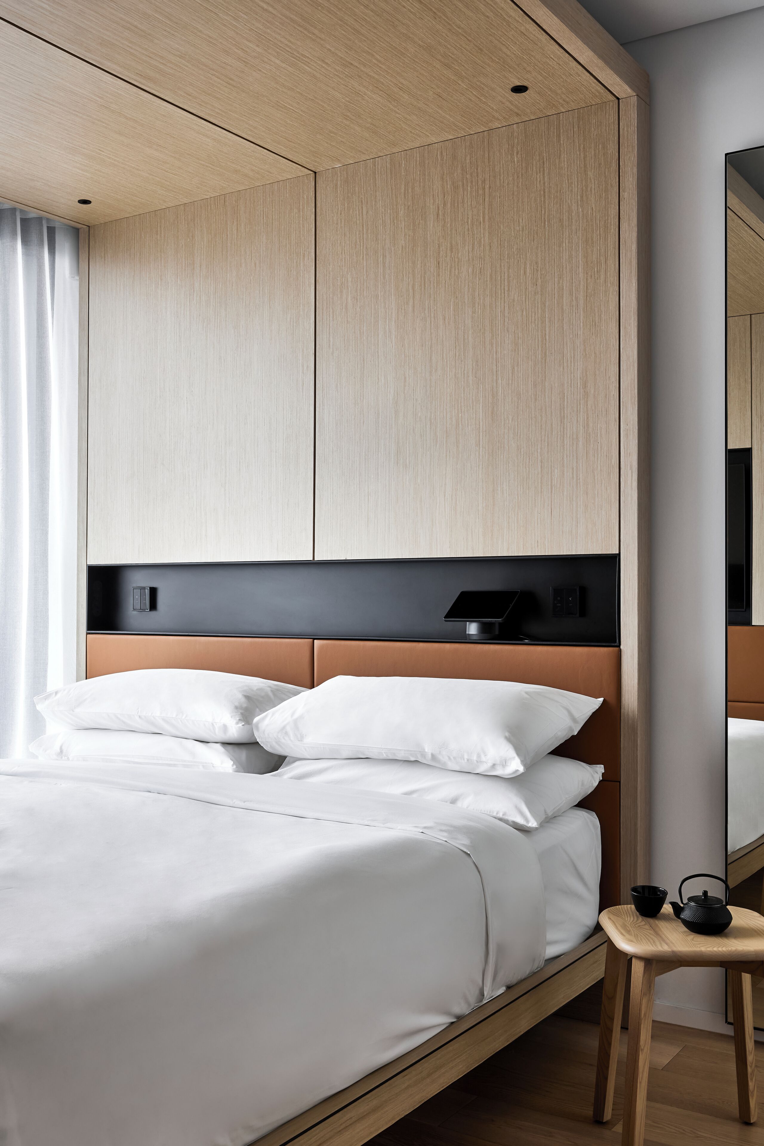 Una camera d'albergo moderna con un letto con struttura in legno, biancheria da letto bianca, un comodino con bollitore nero e un grande specchio a parete.