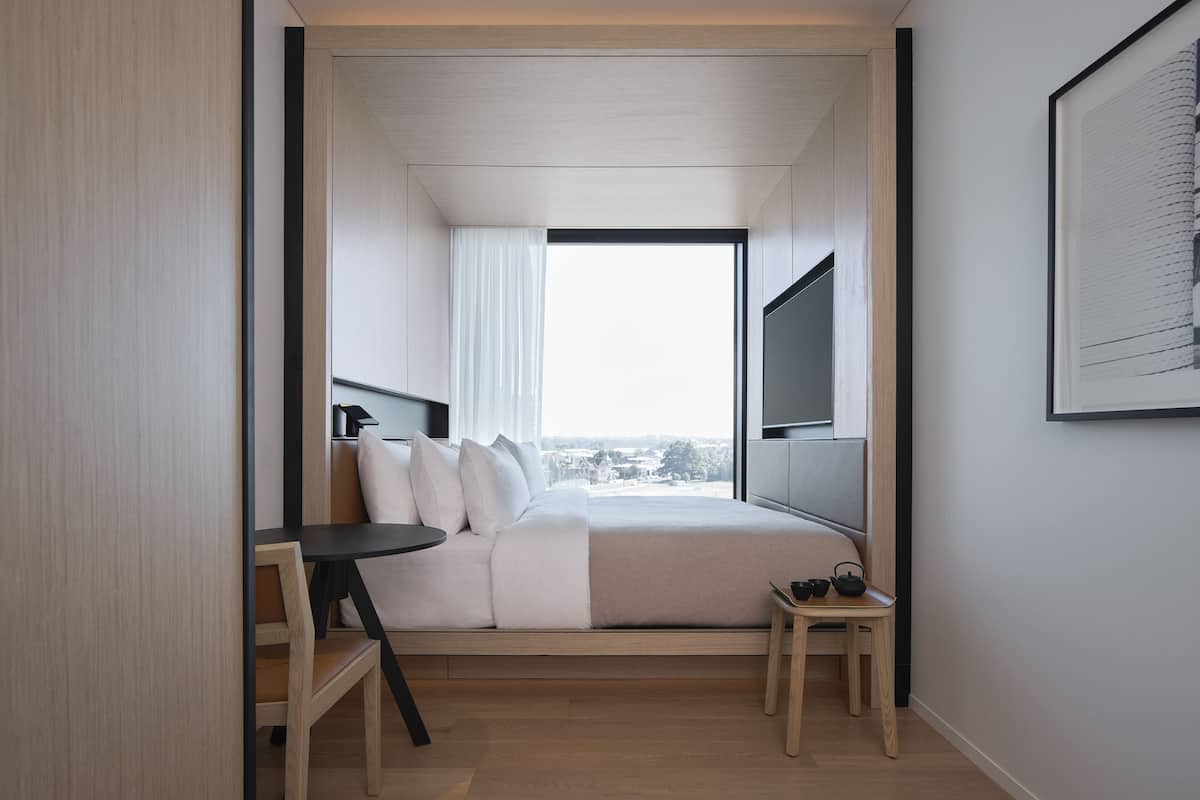A modern hotel room with a bed, a small table, a TV mounted on the wall, and a large window offering a city view.