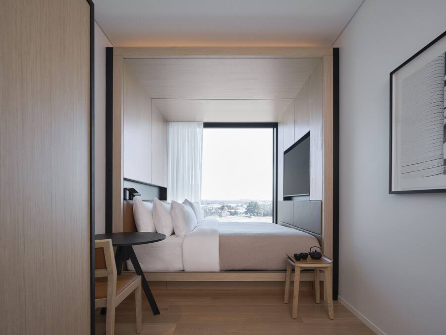 Ein modernes Hotelzimmer mit einem Bett, einem kleinen Tisch, einem an der Wand befestigten Fernseher und einem großen Fenster mit Blick auf die Stadt.