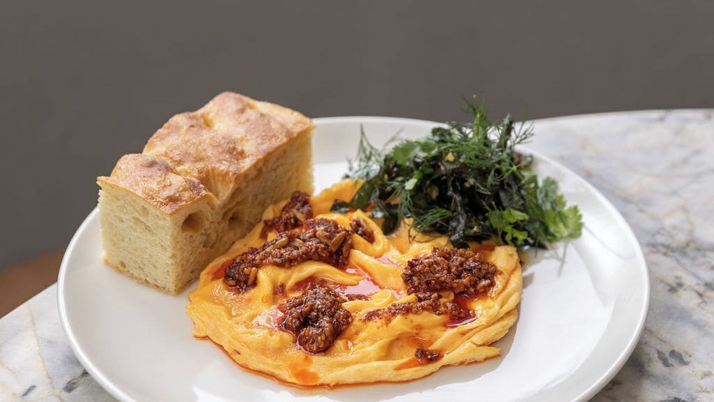 A plate with a dish topped with meat and sauce, accompanied by a piece of bread and a side of greens.