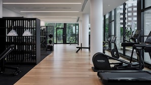 A modern gym with wooden flooring, exercise equipment, and large windows offering views of greenery and buildings.