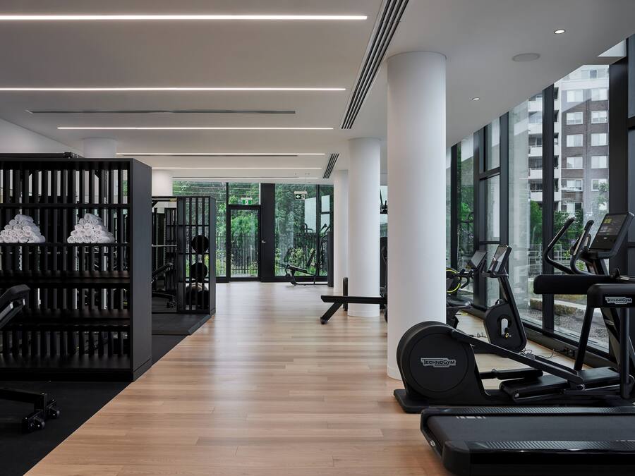 A modern gym with wooden flooring, exercise equipment, and large windows offering views of greenery and buildings.