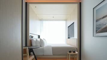 A modern hotel room with a bed, a desk, a chair, a small table with a kettle, and a framed picture on the wall.
