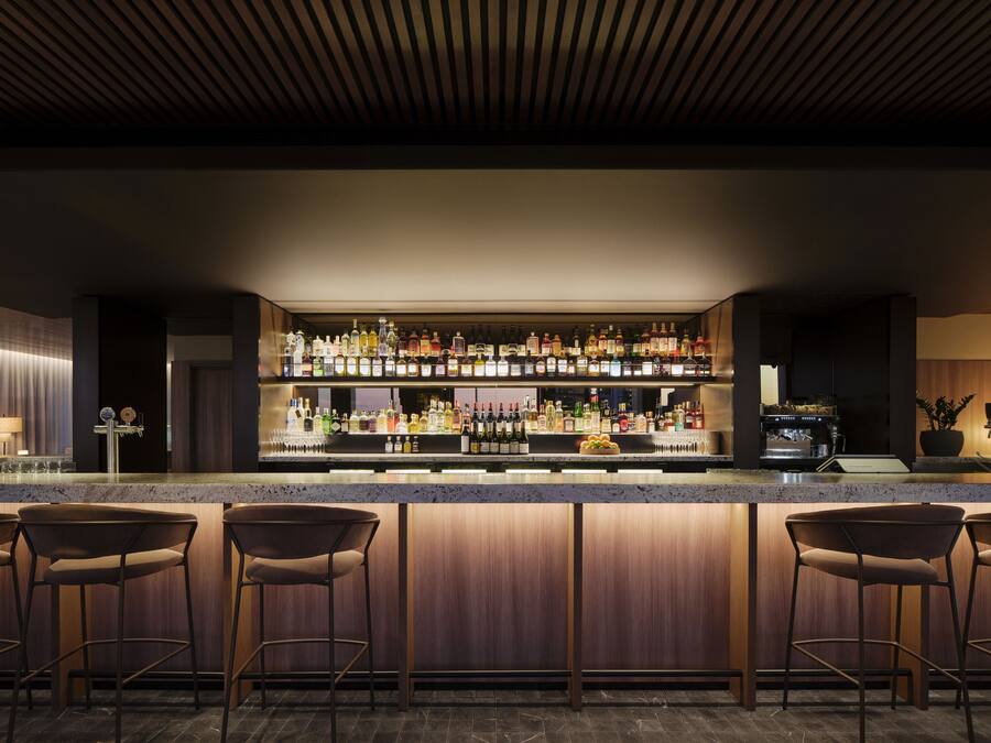 A bar area with a wooden bar counter, multiple shelves of liquor bottles, and three bar stools.