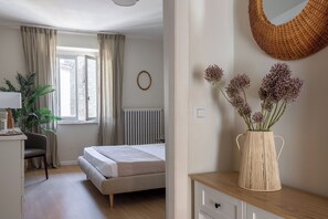 Un dormitorio con cama, un escritorio con silla, una ventana con cortinas, un radiador, un jarrón tejido con flores y un espejo redondo en la pared.