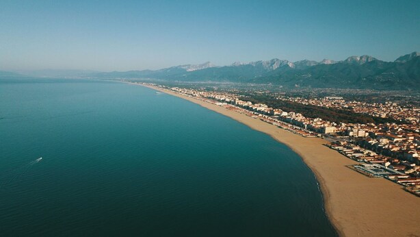 Vue aérienne d’une ville côtière, avec une longue plage, une zone urbaine densément peuplée et des montagnes en arrière-plan.