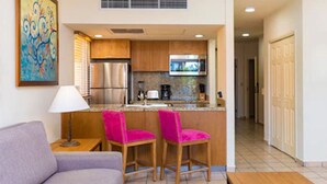A compact kitchen with wooden cabinets, a stainless steel refrigerator, and a microwave. There are two pink bar stools and a small table with a lamp. A painting is on the wall, and a door is visible in the background. - Villa del Palmar (Cabo San Lucas)