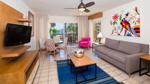 A modern living room with a flat-screen TV, a grey sofa, a pink armchair, a wooden coffee table, and a colorful abstract painting on the wall. - Villa del Palmar (Cabo San Lucas)