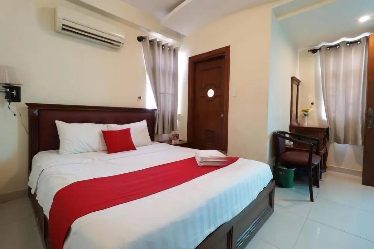 A hotel room with a large bed, a wooden headboard, a desk with a chair, a small table with a vase, and an air conditioning unit on the wall.