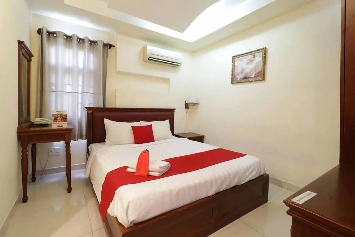 A hotel room with a large bed, a bedside table, an air conditioning unit, a window with curtains, and a framed picture on the wall.