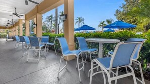 An outdoor patio area with white metal chairs and tables, blue umbrellas, and ceiling fans.