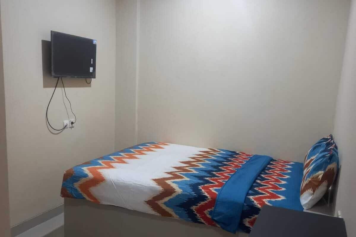Una habitación pequeña y poco amueblada con una cama, un televisor montado en la pared y un escritorio.